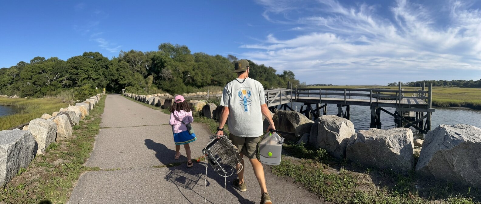 Father and daughter walking to the water carrying lobster traps and a livewell, the man wearing a Checkered Lobster tee